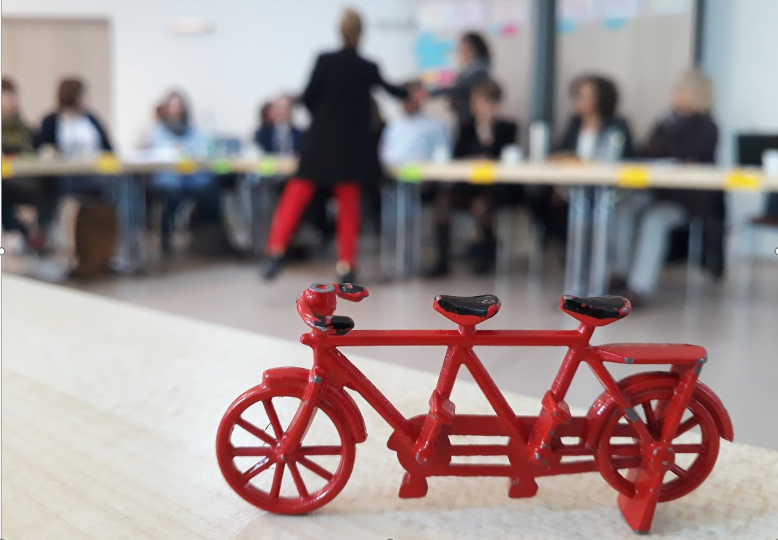Im Vordergrund das Projektsymbol, ein Miniaturtandem, dahinter unscharf Teilnehmende im Workshop