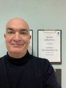 Matthias Süßen Portraitfoto des Journalisten Matthias Süßen. Ein Mann mittleren Alters mit Glatze und schwarzem Rollkragenpullover lächelt in die Kamera. Im Hintergrund steht Willkommen beim Crashkurs Datenvisualisierung