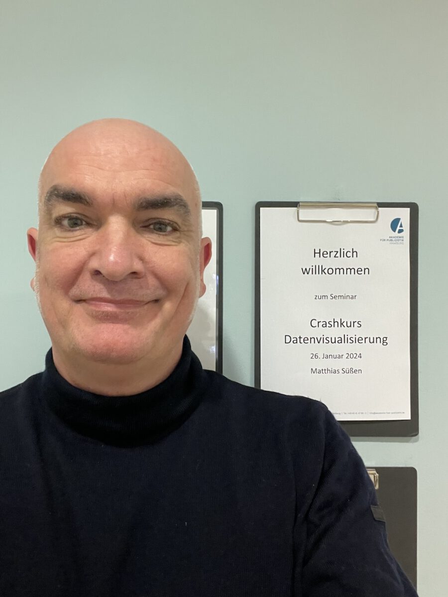 Portraitfoto des Journalisten Matthias Süßen. Ein Mann mittleren Alters mit Glatze und schwarzem Rollkragenpullover lächelt in die Kamera. Im Hintergrund steht Willkommen beim Crashkurs Datenvisualisierung