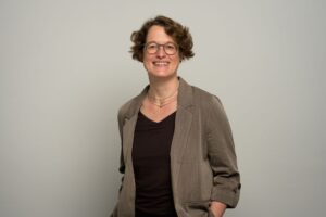 Foto einer Frau mittleren Alters, lächelnd, kürzere krause Haare, mit brauner Jacke und schwarzem Oberteil